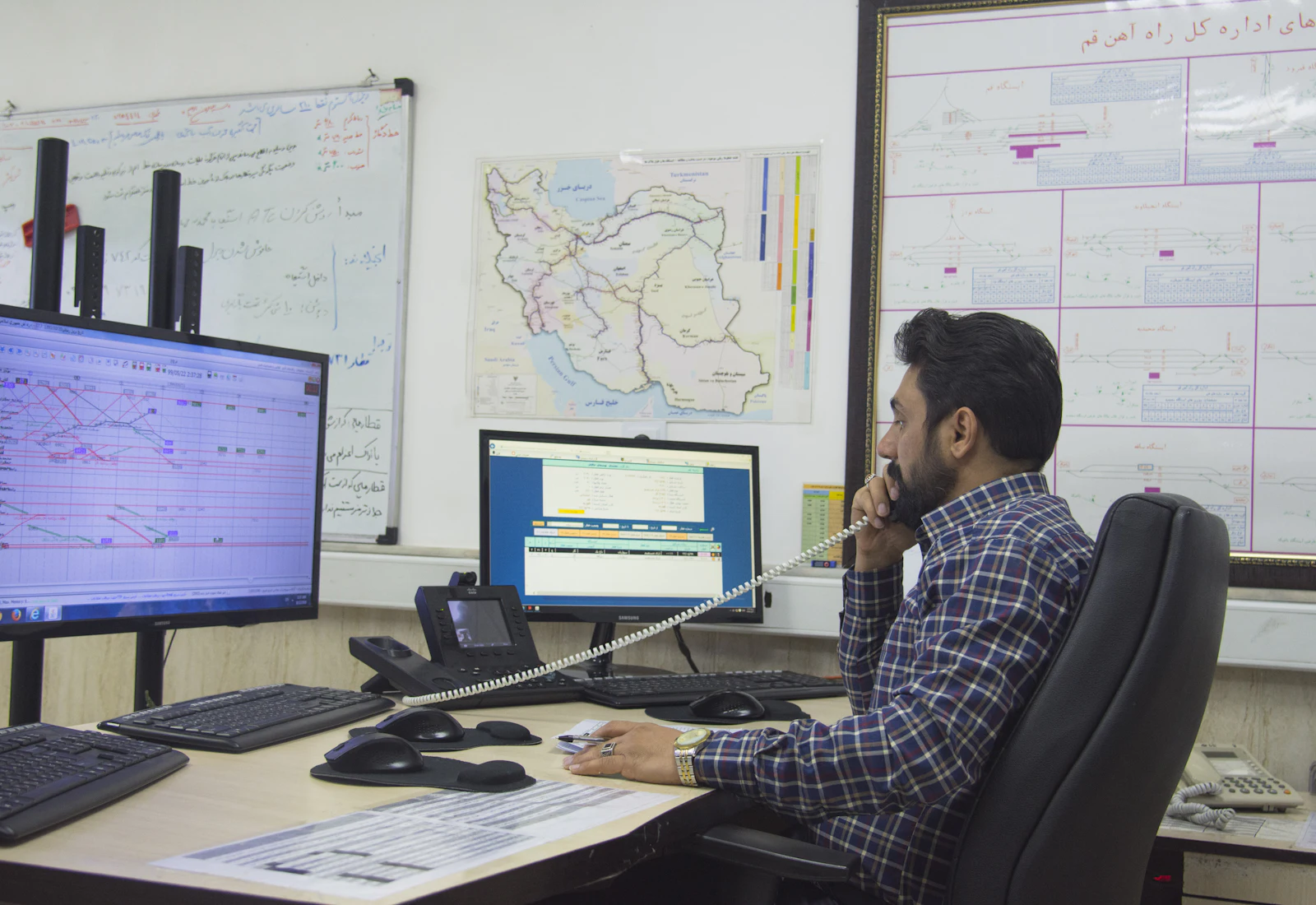 A dispatcher on the phone at a desk, working from wall planners and a spreadsheet.