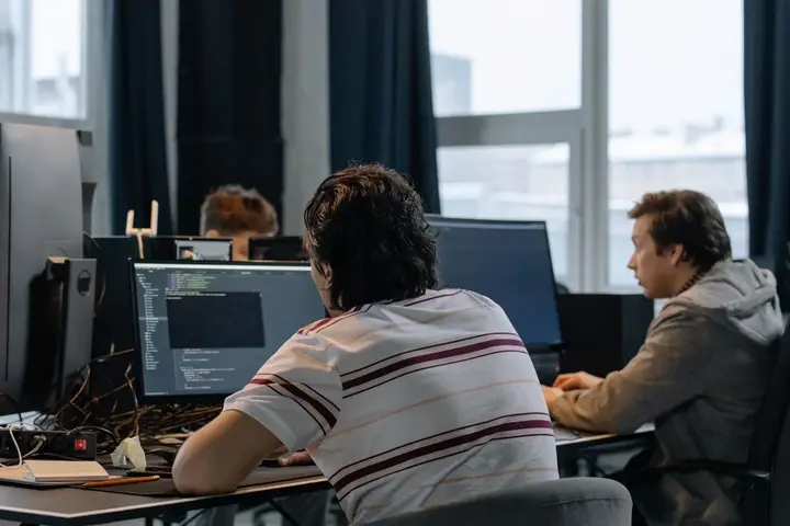 Developers at shared desks in a bright studio, one reading code on a large monitor.