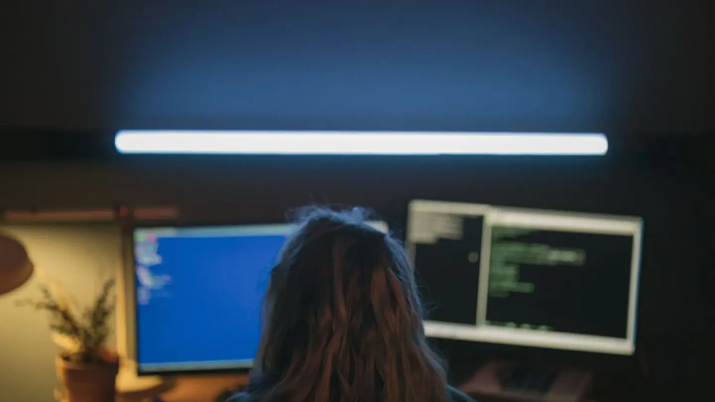 A developer seen from behind at night, working across two monitors showing code under a strip light.