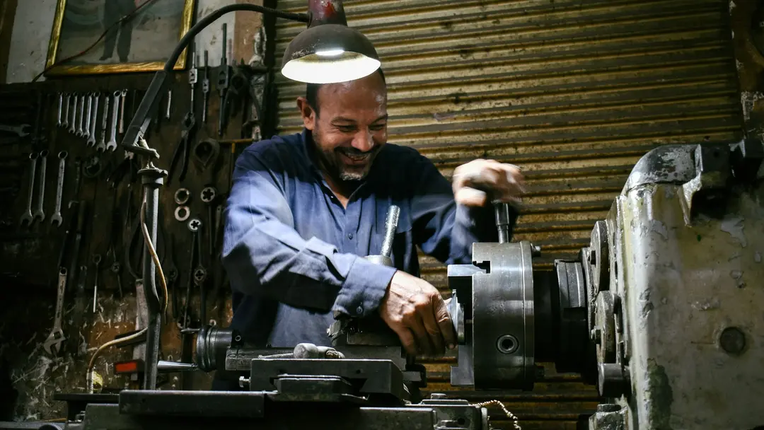 A machinist at a lathe in a workshop, hand on the cross-slide, tools racked on the wall behind him.