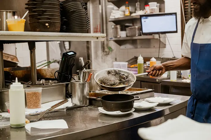 A restaurant pass during service, a ticket screen on the wall behind it and a cook lifting a pan of shellfish.