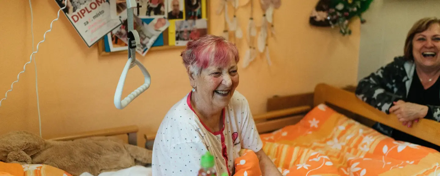 A woman with pink-streaked hair laughing in a bright room, another woman smiling behind her.