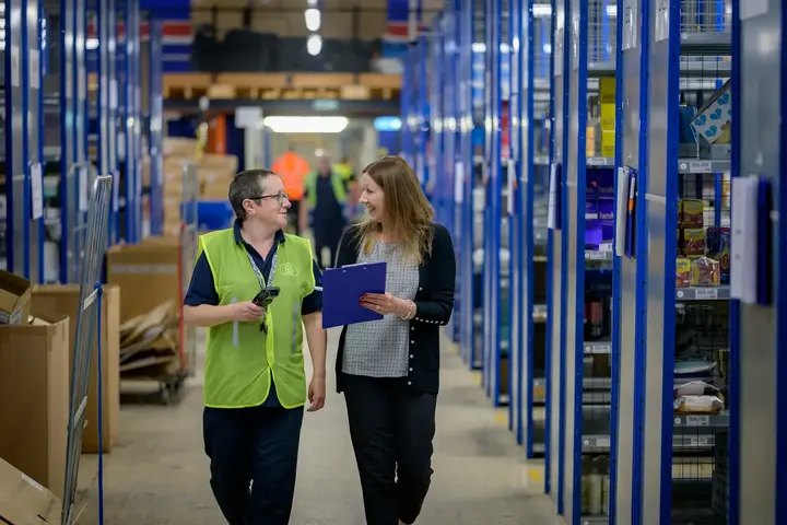 Two colleagues in hi-vis walking a warehouse aisle, one holding a clipboard.