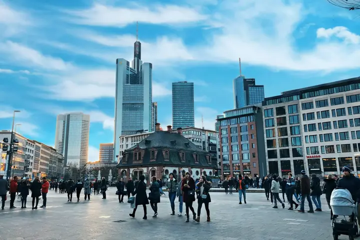 People crossing a wide paved square in central Frankfurt, with the banking district’s towers rising behind.