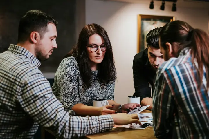 Four colleagues leaning over a table together, passing printed pages between them.