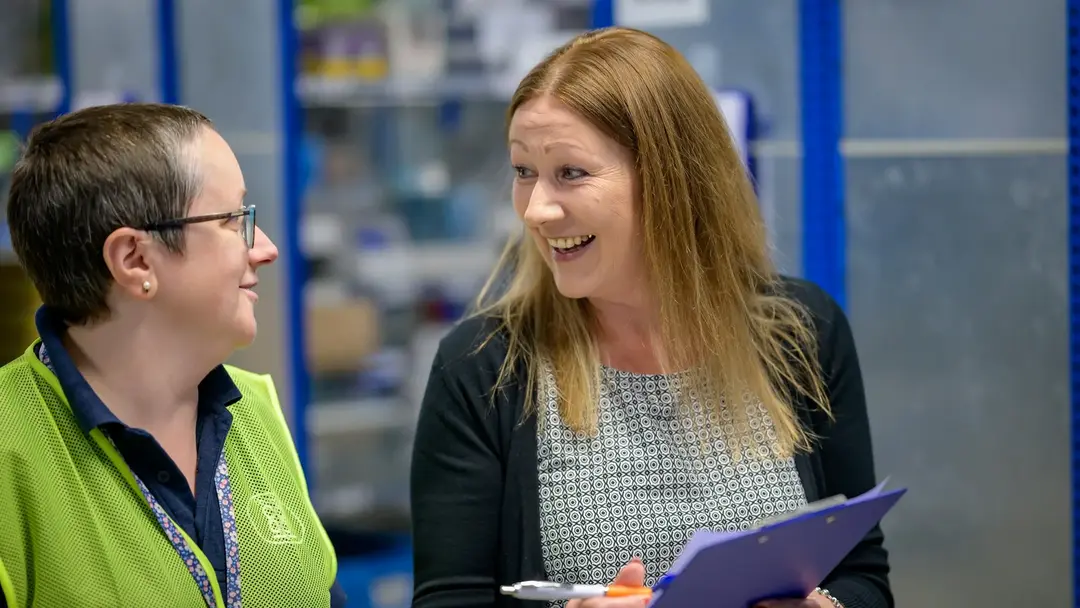 Two women in a warehouse stockroom mid-conversation, one in a work polo and lanyard, the other holding a clipboard.
