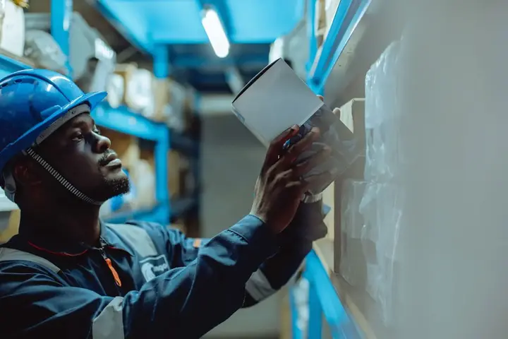 A worker in a hard hat holding a tablet against a warehouse rack, checking a label.