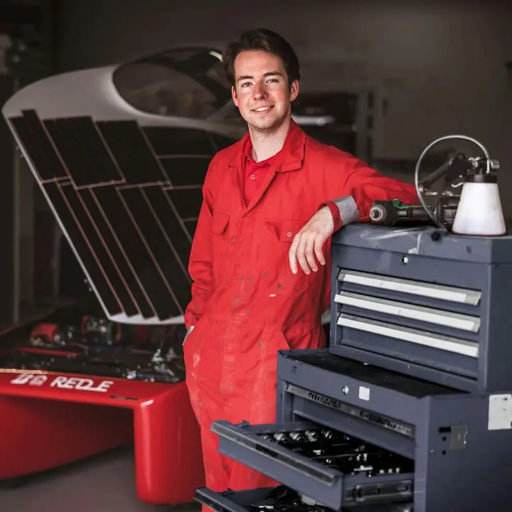 A mechanic in red overalls standing beside his tool chest, smiling.