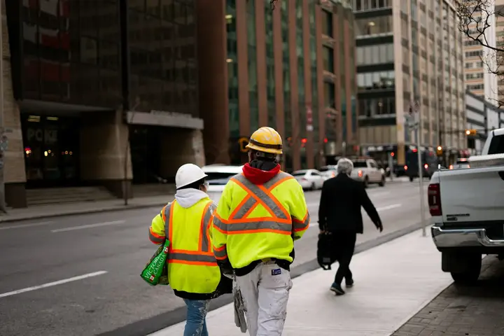 Two workers in high-visibility jackets walking along a city street between office blocks.