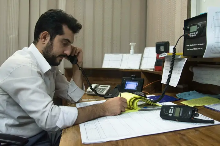 A man at a cluttered desk with a phone to his ear, writing in a ledger, a two-way radio and a base station beside him.