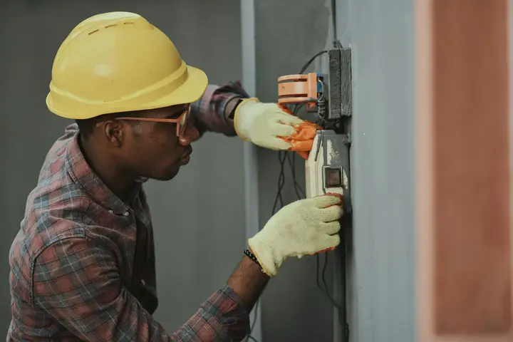 An electrician in a hard hat and work gloves fitting a meter box to a wall.