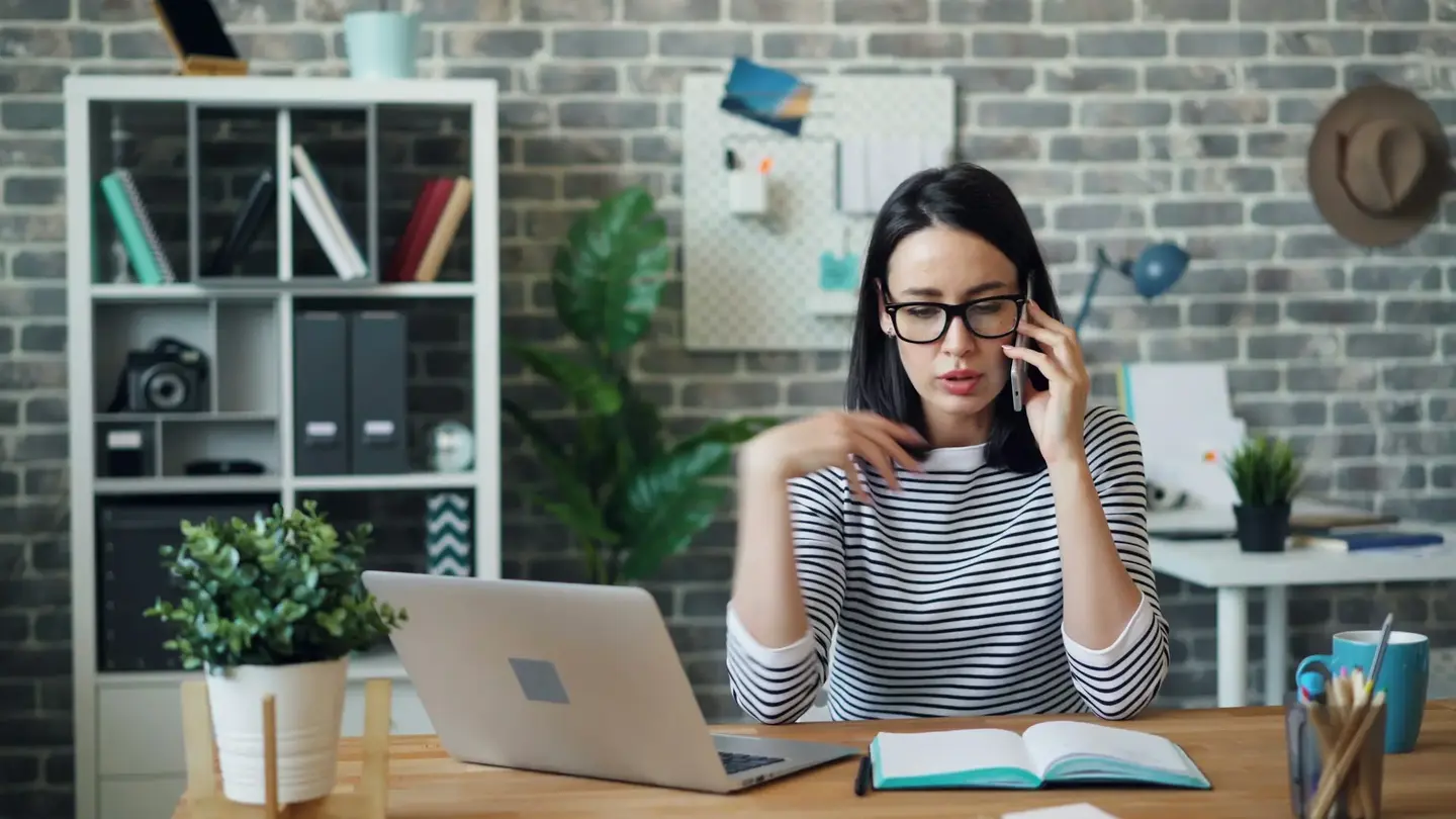 A woman at a desk with a laptop and an open notebook, on the phone, frowning slightly as she listens.