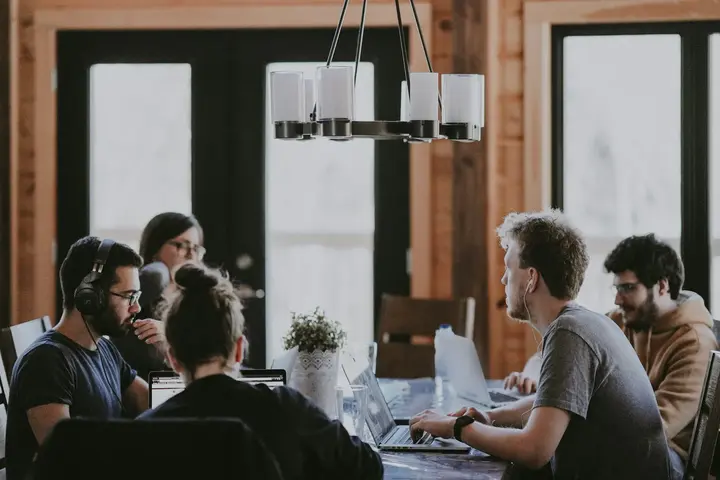 Five people working on laptops around a long wooden table in a bright room, one in headphones.