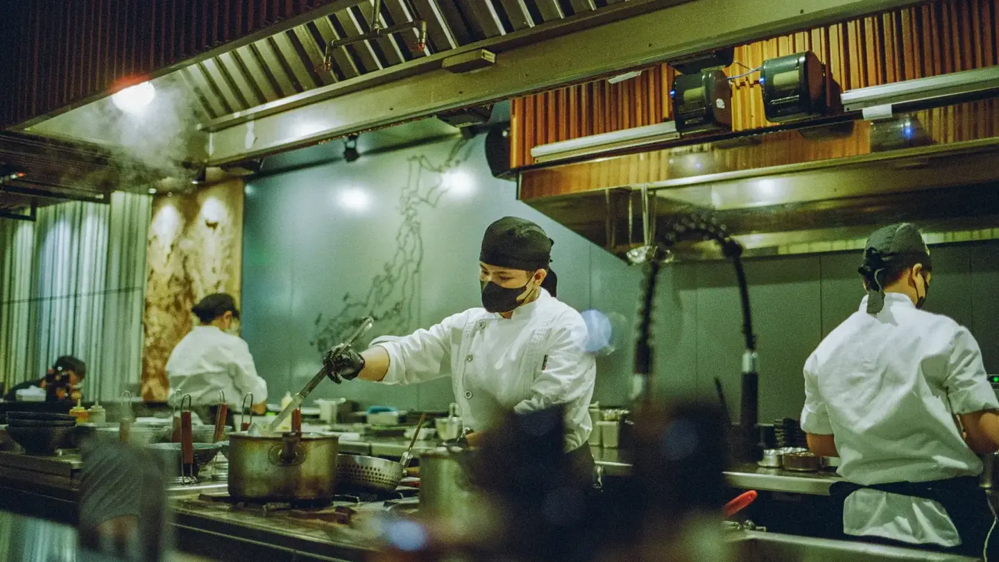 Three cooks working the line in a restaurant kitchen during service, ticket printers mounted above the pass.