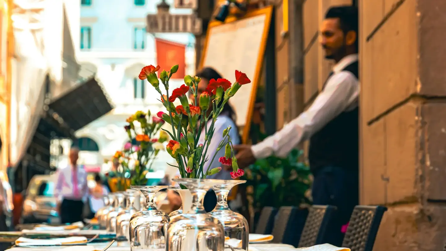 A waiter at a restaurant door beside a menu board, the terrace tables laid with glasses and flowers before service.