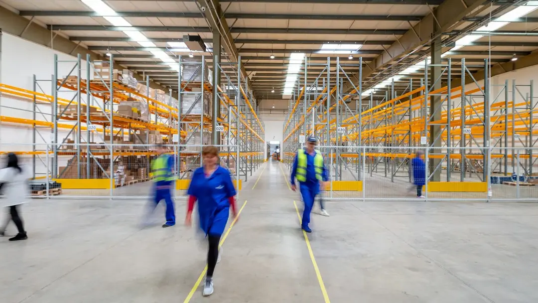 A wide warehouse aisle at shift change, several workers walking through it, caught mid-stride.