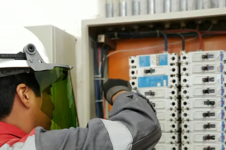 An electrician in a face shield testing terminals inside a distribution board.
