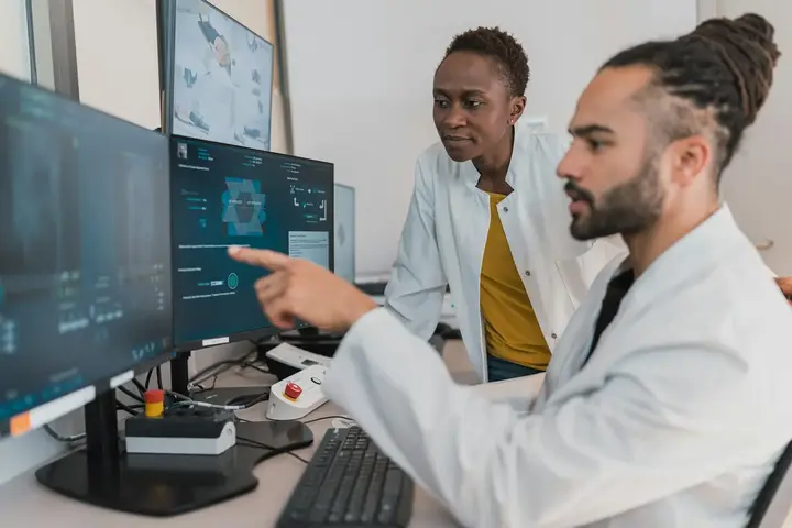 Two clinicians in white coats reading a scan together at a workstation.