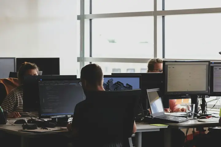 A support floor from behind: people at paired screens with headsets on the desks, backlit by a window.