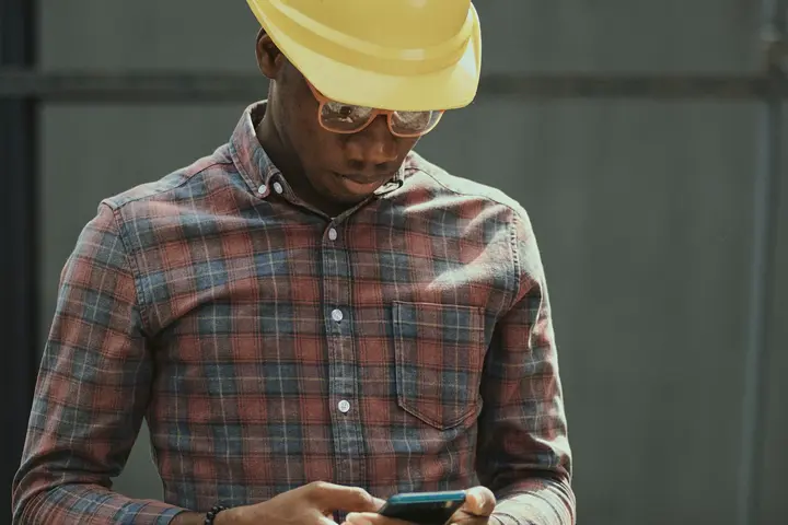 A worker in a hard hat and safety glasses on a site, reading something on the phone in both hands.