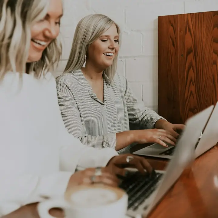 Two women laugh as they work on laptops side by side at a café counter.