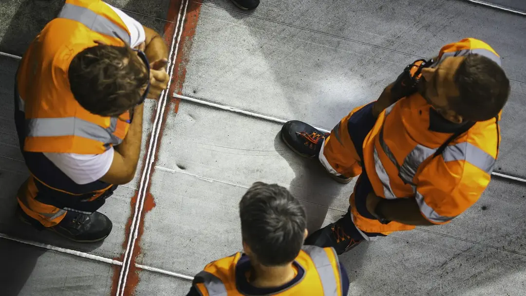 Three crew in high-visibility kit waiting on a vehicle deck, one of them on a radio, seen from above.