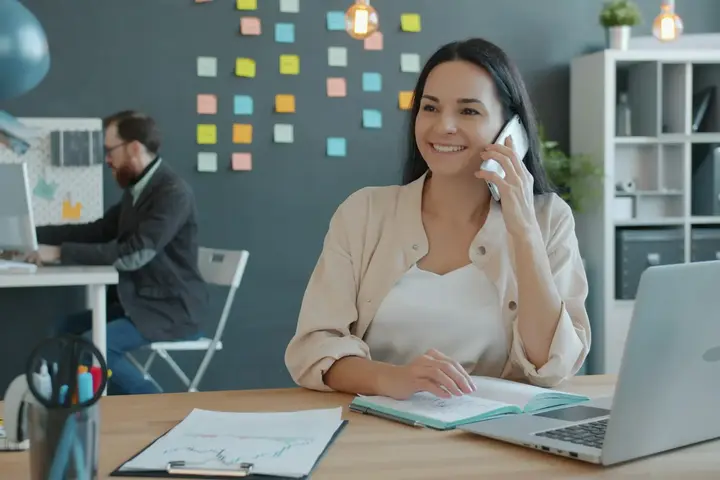 A woman smiles while on a phone call at her desk, with a laptop and a notebook in front of her.