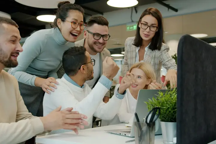 A group of colleagues cheer and laugh together as they look at a laptop screen.