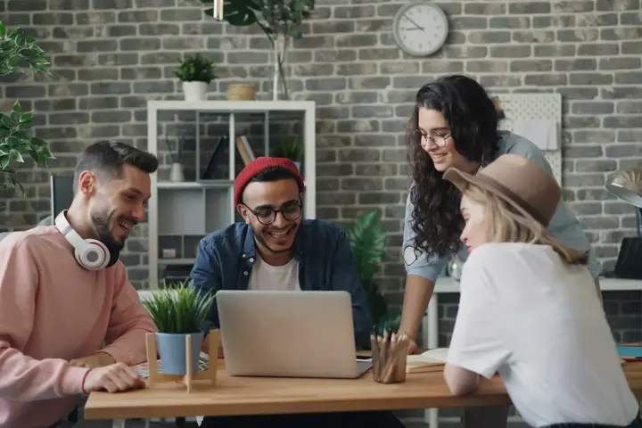 Four young colleagues laugh together around a laptop in a brick-walled office.
