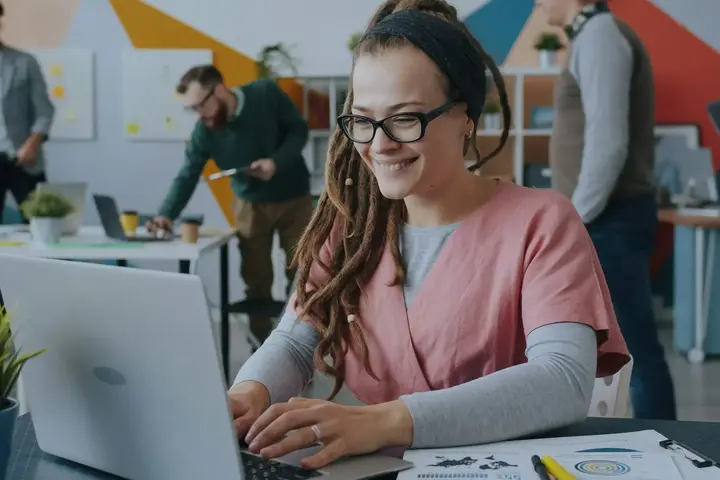 A woman in glasses smiles as she types on a laptop in a busy, colourful office.