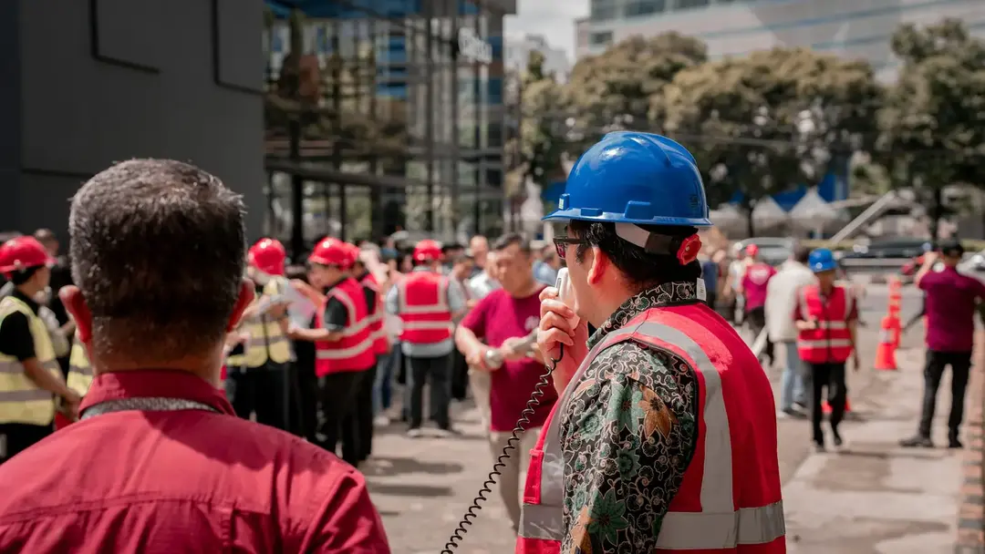 A site foreman speaking into a handheld radio, with a crew in hi-vis assembled behind him.