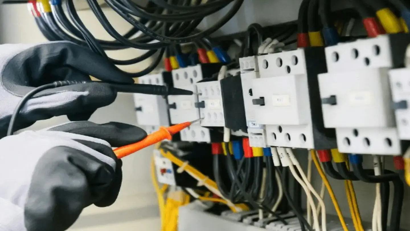A gloved electrician testing terminals inside a consumer unit with a multimeter probe.