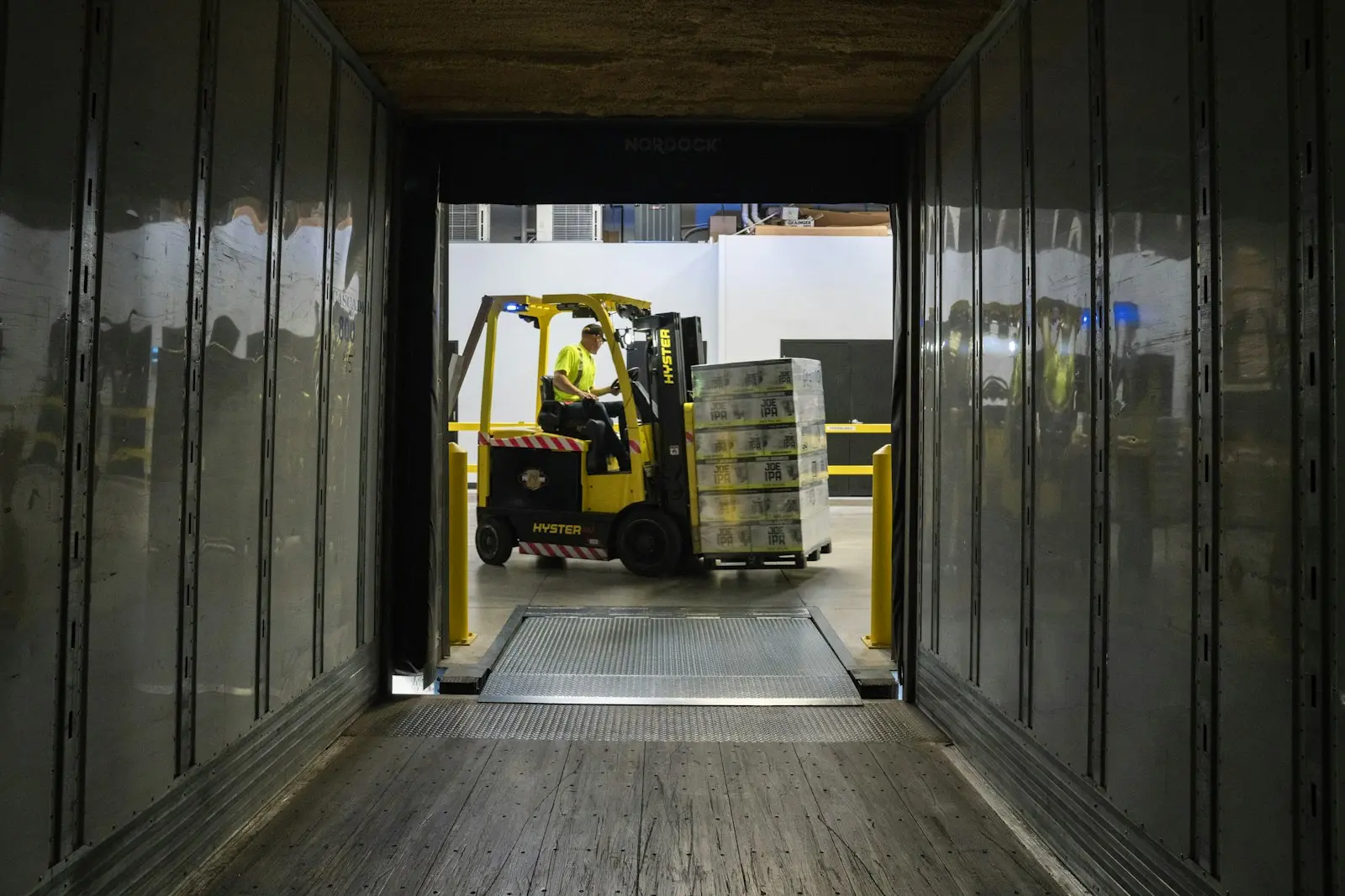 A forklift carrying a pallet up the ramp into a trailer, seen from inside the trailer.